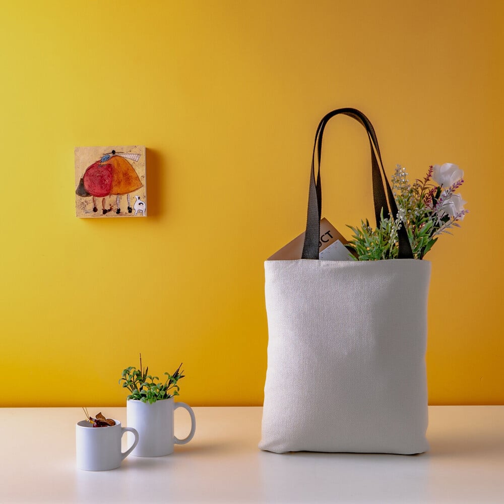 A tote bag with flowers and a box in it - next to the tote bag, there are two coffee cups with plants in them and a child's drawing hanging on the wall.