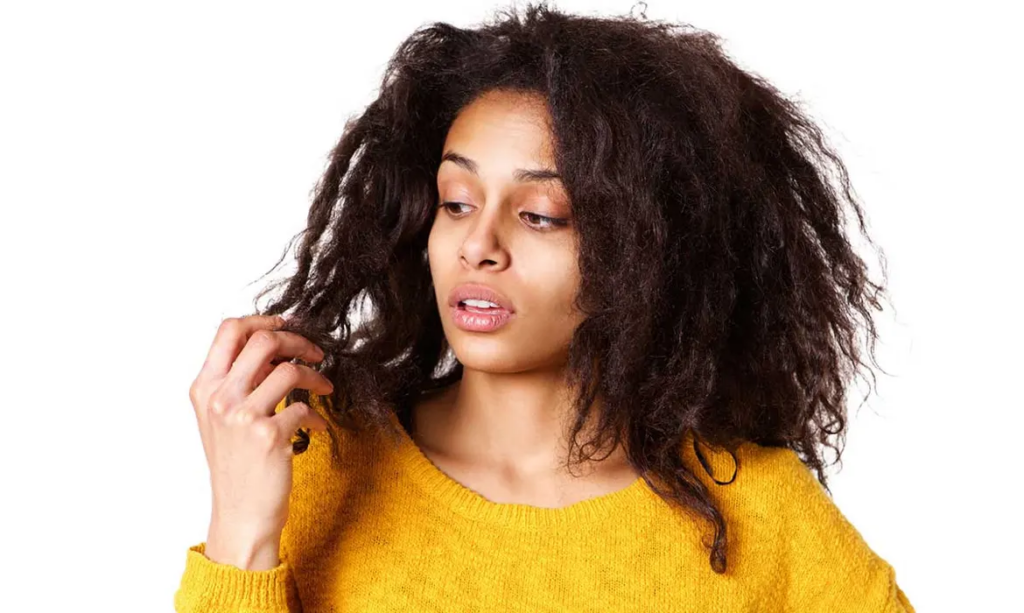Get the Facts About Chemical Relaxers and Hair Loss - a woman in a yellow sweater inspecting her chemically damaged hair with her right hand.