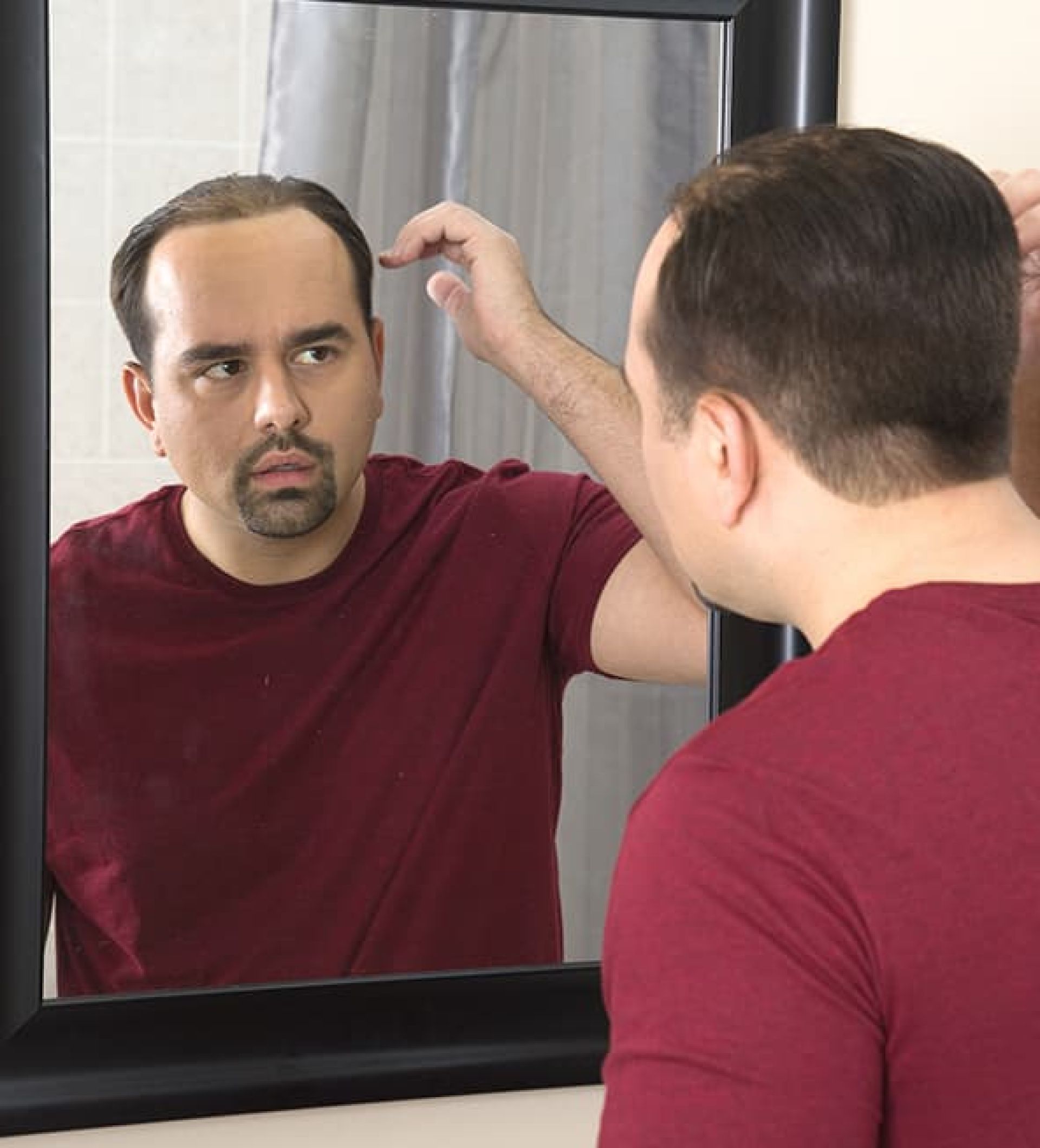 HairClub Causes and Conditions - a man wearing a burgundy t-shirt looks at his male pattern baldness receding hairline in a mirror.