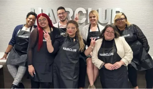 HairClub - About - Join Us - 7 diverse HairClub employees posing for a picture infront of the reception desk at a HairClub center - 2 are making peace signs - the HairClub logo is hanging on the wall behind them.