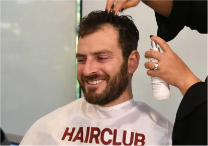 HairClub - EXT Max Results - a man getting an Extreme Hair Therapy product spayed on his hair by a HairClub Hair Loss Specialist - he is wearing a barber's cape with the HairClub logo on it.