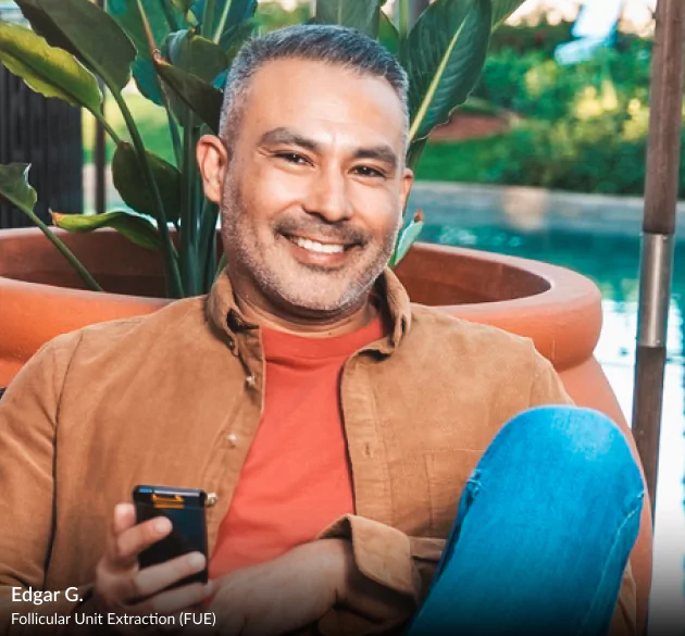 HairClub - Our Solutions - a man sitting outside by a pool - he is smiling and wearing a red t-shirt underneath an open brown button-down - he has salt and pepper hair and a perfectly straight hairline.