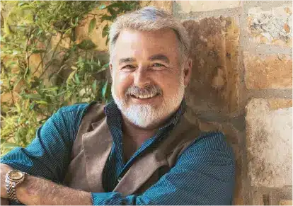 HairClub - Learn Men- a bearded older gentleman with a nice head of hair is wearing a blue striped button-up shirt and a brown vest - he's smiling and standing next to a stone wall and a green plant.