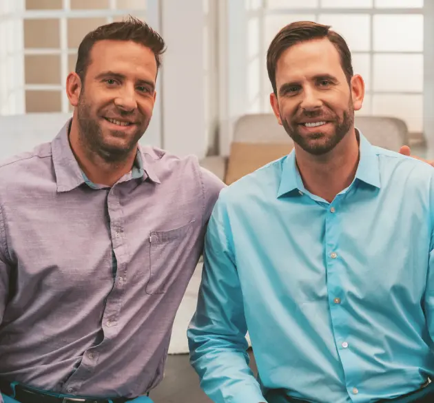 HairClub - Male Pattern Hair Loss - two brothers - the one in a purple dress shirt has his arm around the the other in a teal dress shirt as they pose for a photo.