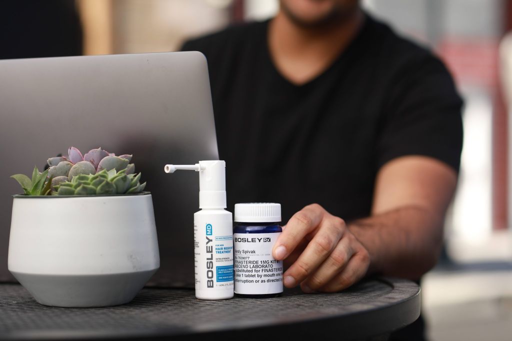 A close-up of a small table with a laptop and two Bosley hair treatment products - a spray bottle Bosley MD and a pill bottle Bosley Rx