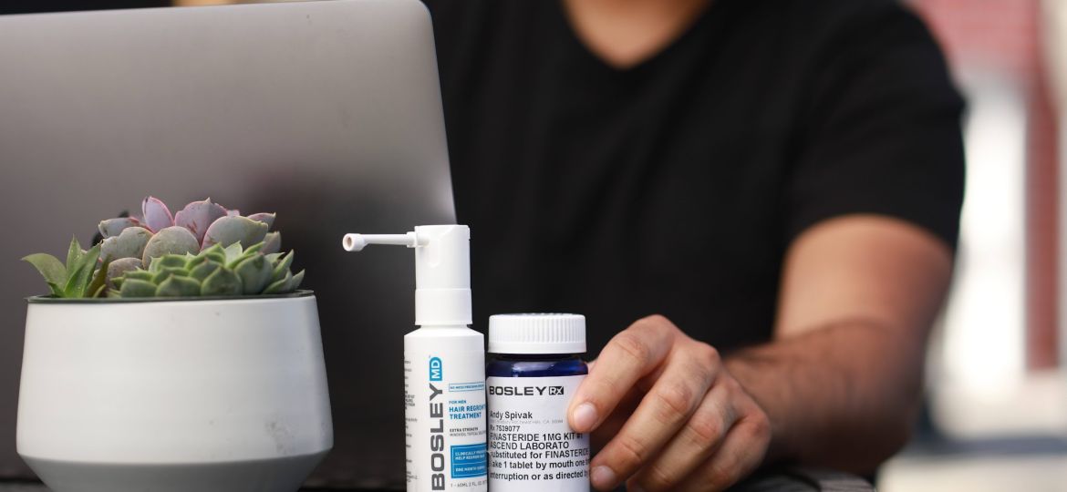 A close-up of a small table with a laptop and two Bosley hair treatment products - a spray bottle Bosley MD and a pill bottle Bosley Rx