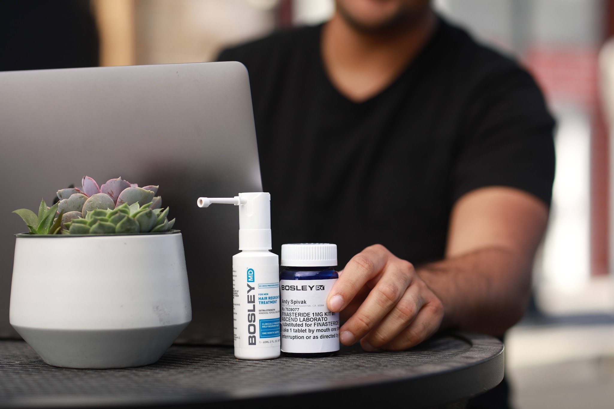 A close-up of a small table with a laptop and two Bosley hair treatment products - a spray bottle Bosley MD and a pill bottle Bosley Rx
