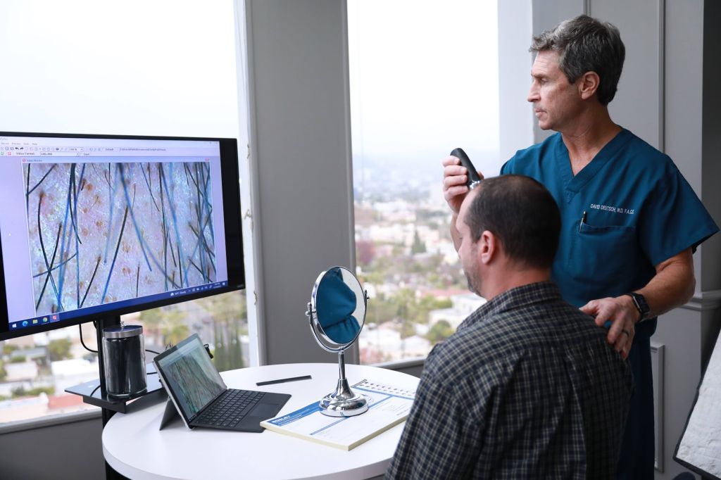A doctor in blue scrubs is examining a male patient's scalp using handheld dermatoscope in a medical office with large windows.