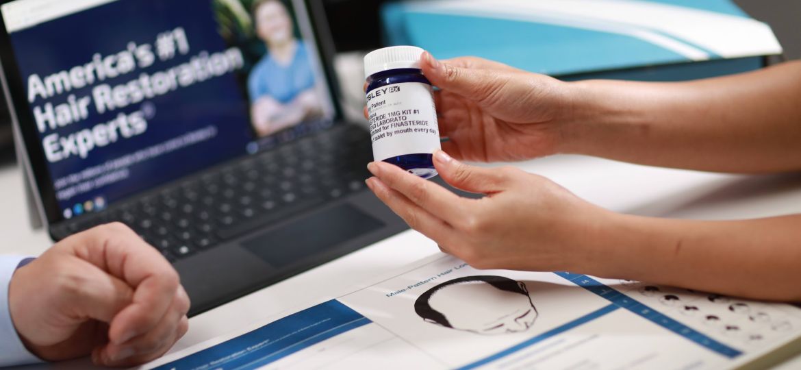 A person holds a Finasteride bottle in front of a laptop displaying the bosley website
