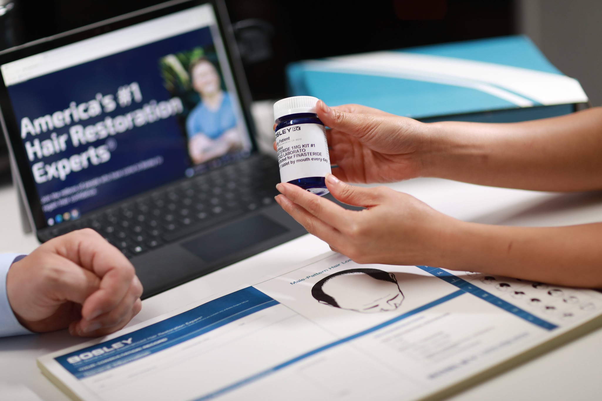 A person holds a Finasteride bottle in front of a laptop displaying the bosley website