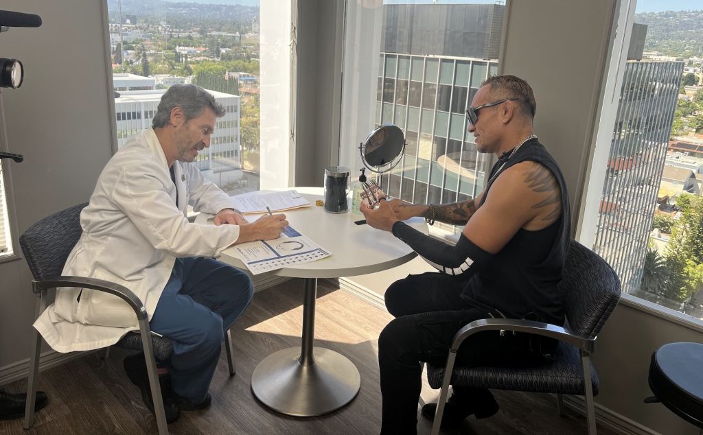 Hair restoration consultation in Bosley Beverly Hills clinic, showing a doctor reviewing paperwork while a male patient examines hair samples at a small table with city skyline views through large windows.