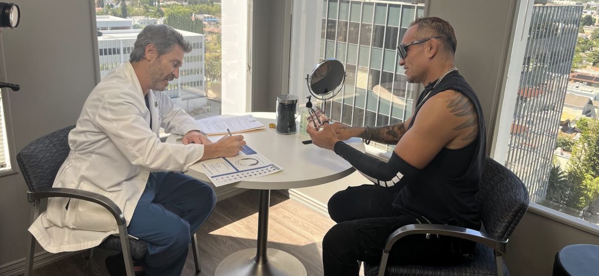 Hair restoration consultation in Bosley Beverly Hills clinic, showing a doctor reviewing paperwork while a male patient examines hair samples at a small table with city skyline views through large windows.