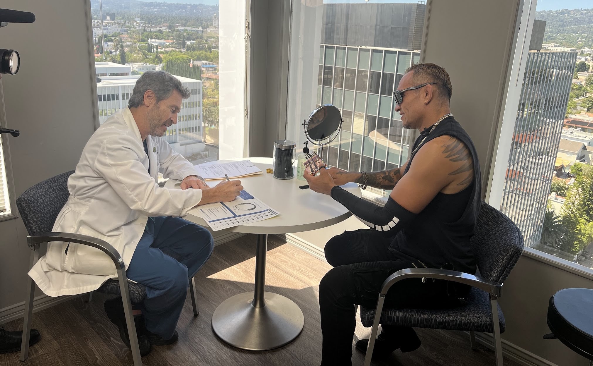 Hair restoration consultation in Bosley Beverly Hills clinic, showing a doctor reviewing paperwork while a male patient examines hair samples at a small table with city skyline views through large windows.