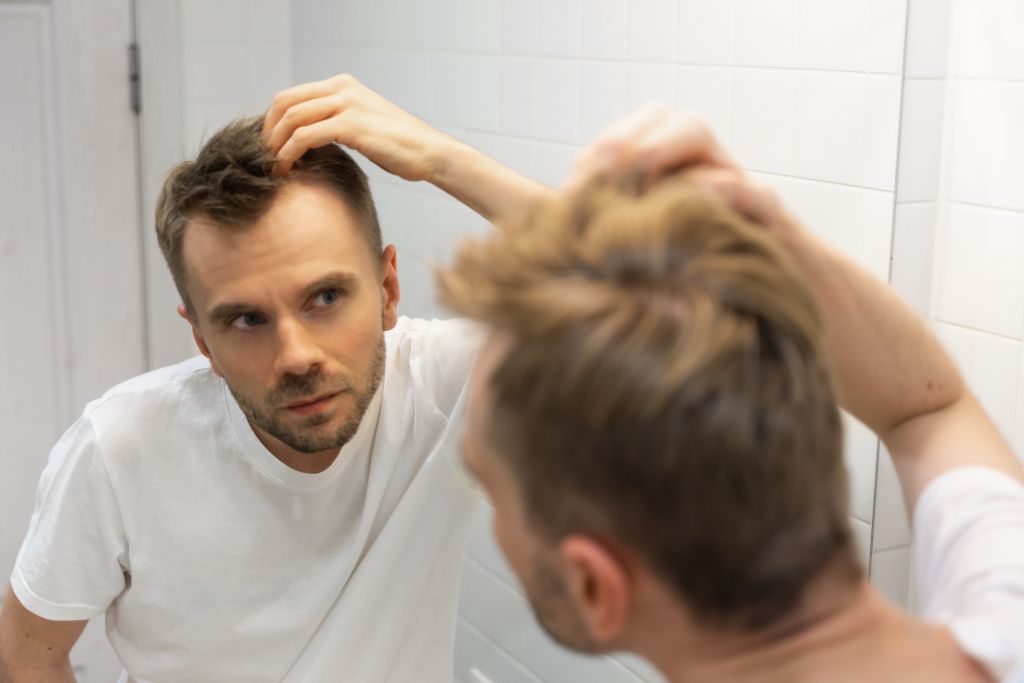 Middle aged caucasian white man with a short beard looks at his hair in the mirror in the bathroom and worried about balding. The concept of the problem of male hair loss, early baldness and alopecia.